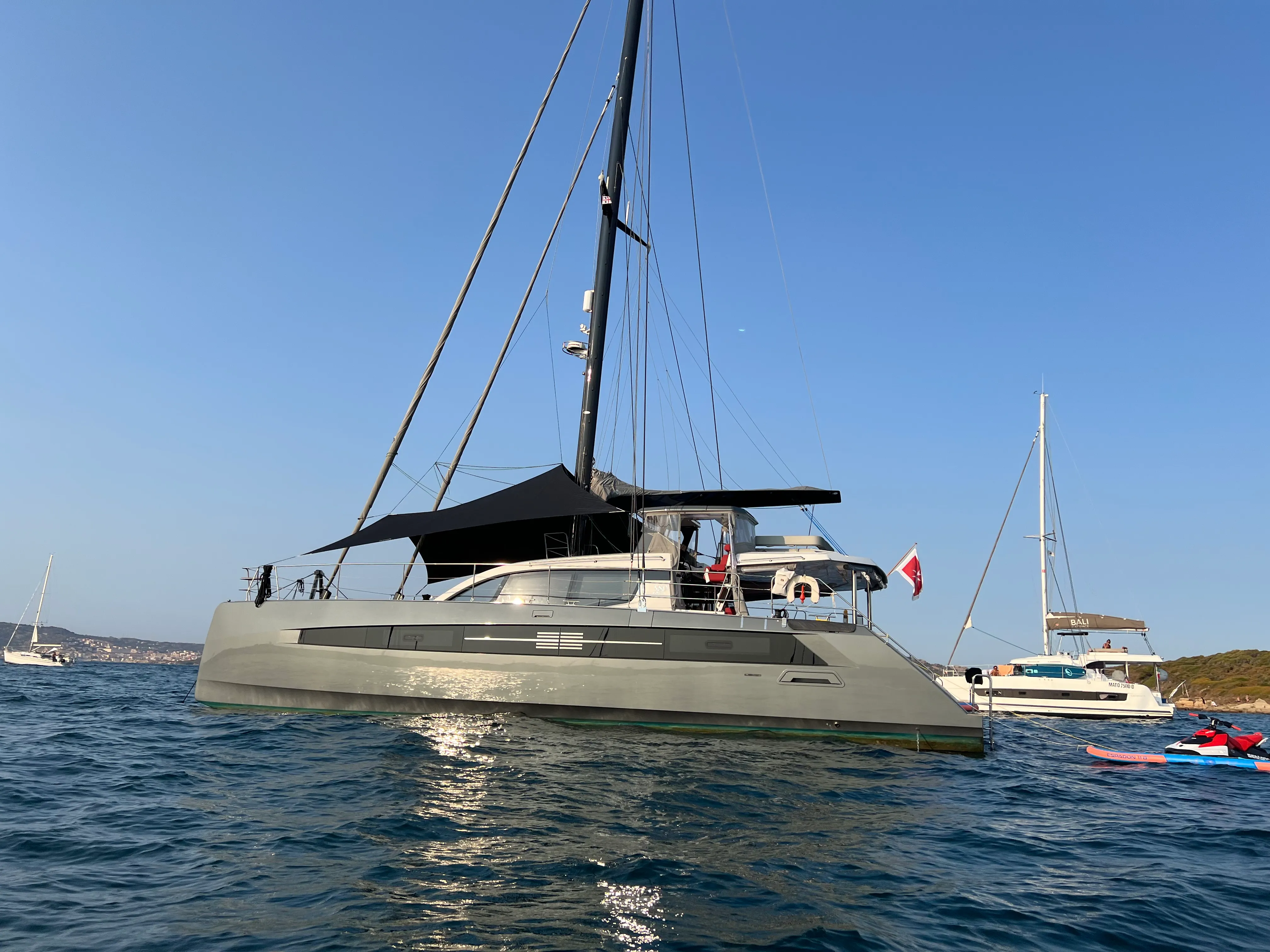 Mymlan, a dark-hulled Privilège sailing catamaran at anchor, with paddleboard and jet ski alongside